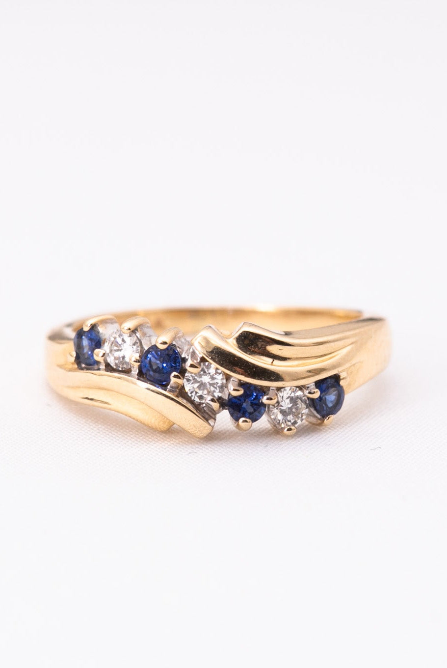 A gold ring featuring a wave-like design with small blue sapphires and clear round-cut stones, photographed on a plain white background.