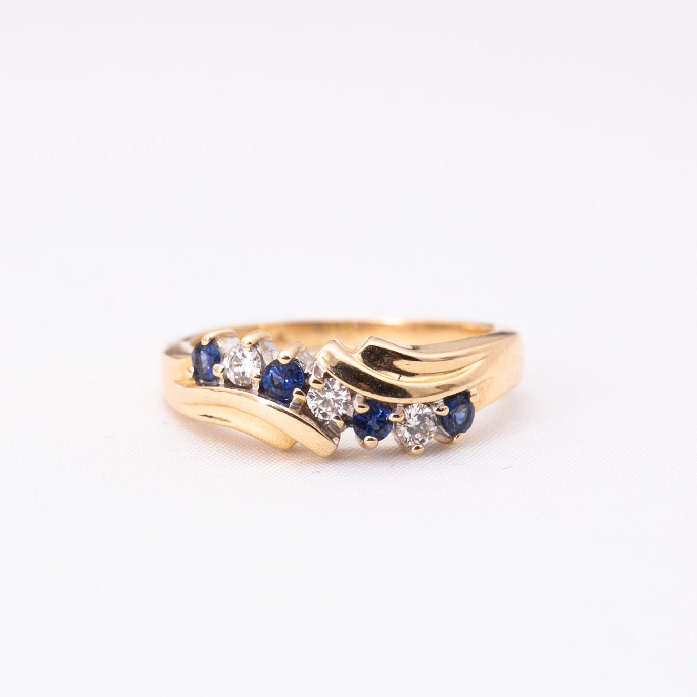A gold ring featuring a wave-like design with small blue sapphires and clear round-cut stones, photographed on a plain white background.