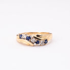 A gold ring featuring a wave-like design with small blue sapphires and clear round-cut stones, photographed on a plain white background.